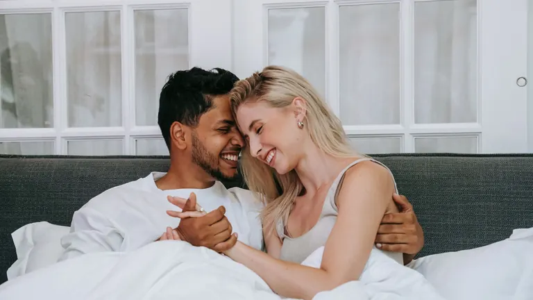 Smějící se pár v posteli pod bílými peřinami při intimní chvíli